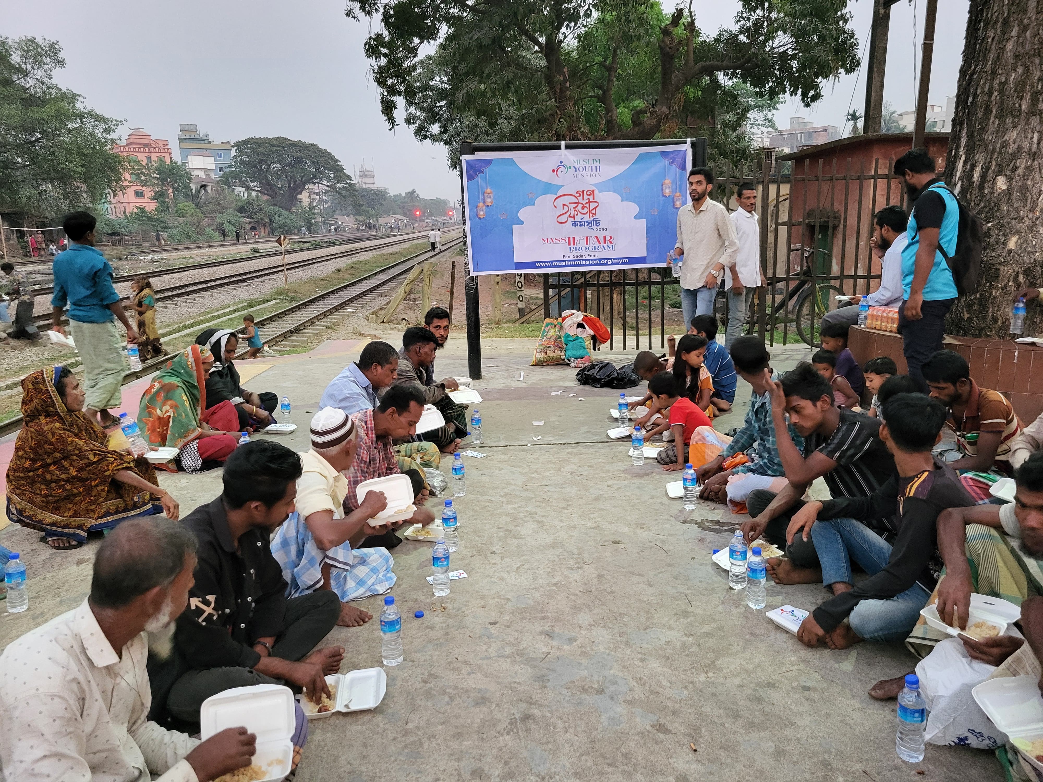 A group of people, mostly men, are seated on the ground near a railway track, with a large banner visible in the background.
