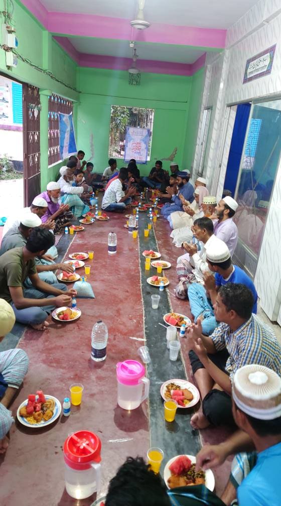 A large group of men are seated on the floor inside a mosque, eating together in a brightly colored room.