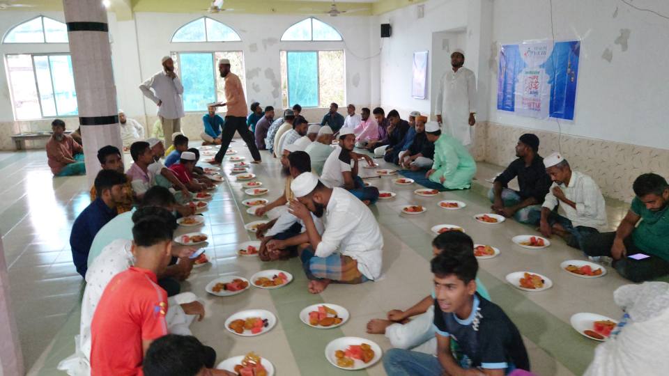 A large group of people, mostly seated, are gathered in a spacious room with arched windows, engaged in what appears to be a communal meal or gathering.