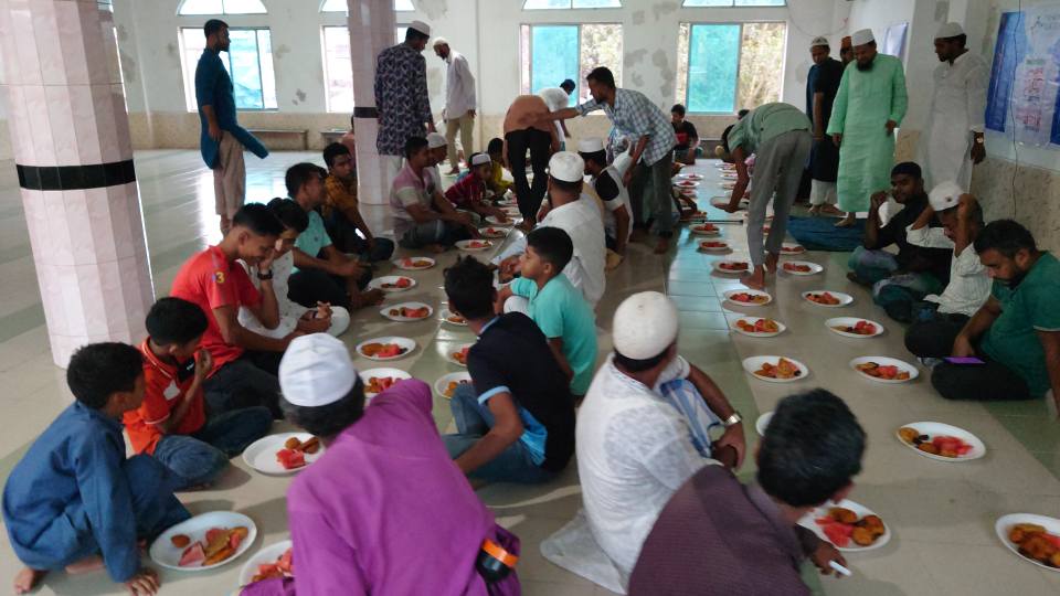 A large group of people, both adults and children, are seated at tables in a spacious room, enjoying a communal meal together.
