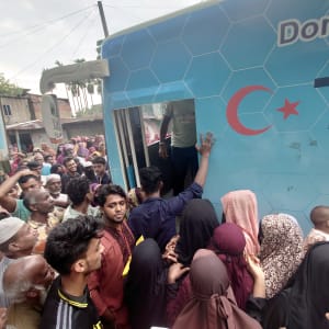 A crowd of people surrounds a blue vehicle with a red star, likely in an urban setting.