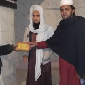 Three men in traditional attire exchange a gift in a white-tiled room.