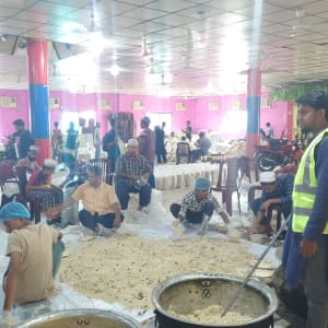 Crowd of people eating from large pots in a hall, likely a communal meal.