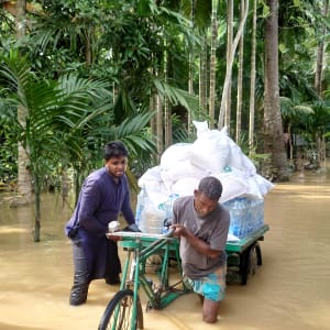 Two men push a green flatbed cargo tricycle loaded with sacks through floodwater in a tropical forest.
