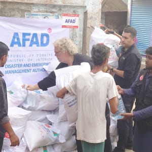 Man in a blue uniform holding a bag of supplies, likely aid distribution, against a gray wall.