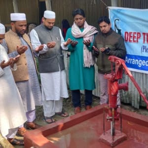 Several men are praying beside a water pump, likely in a rural setting.