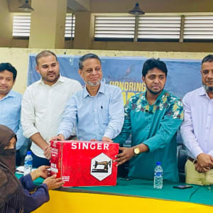 Several men are standing behind a table presenting a Singer sewing machine box to a woman wearing a black burqa, likely at a meeting.