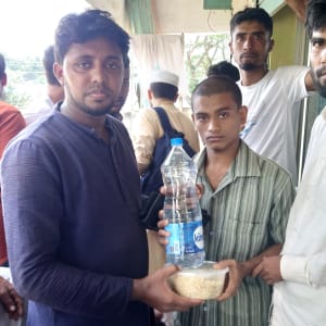 A young man offers a bag of rice to a group of men in a line, likely during a humanitarian aid distribution.