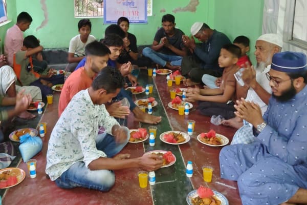 A group of people, mostly men, are gathered together in a colorful, open-air setting, sharing a meal and engaging in conversation.
