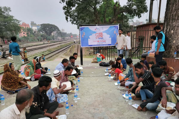 A group of people, mostly men, are seated on the ground near a railway track, with a large banner visible in the background.