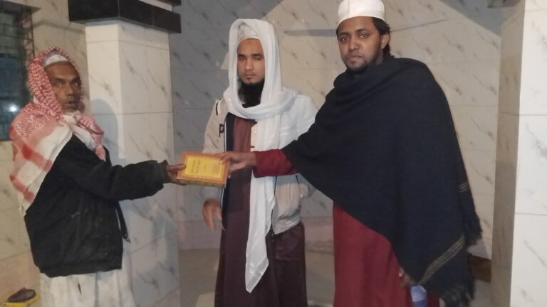 Three men in traditional Islamic dress standing together indoors. The man on the right wears a black and maroon robe, the man in the middle wears white robes with a head covering, and the man on the left wears a red and white checkered headdress. They are holding what appears to be a yellow book between them.