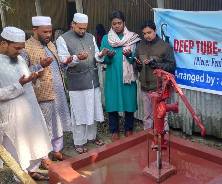A group of five men stand around a red hand-pump water well, raising their hands in prayer. A banner in the background reads "Deep Tube-Well" with additional text about its location and organisers.