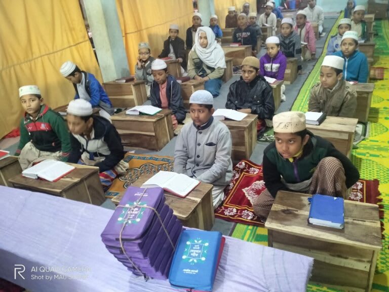A classroom filled with children, seated on the floor with wooden desks in front of them. They are dressed in warm clothing and wearing white caps, studying religious texts in a madrasa setting.