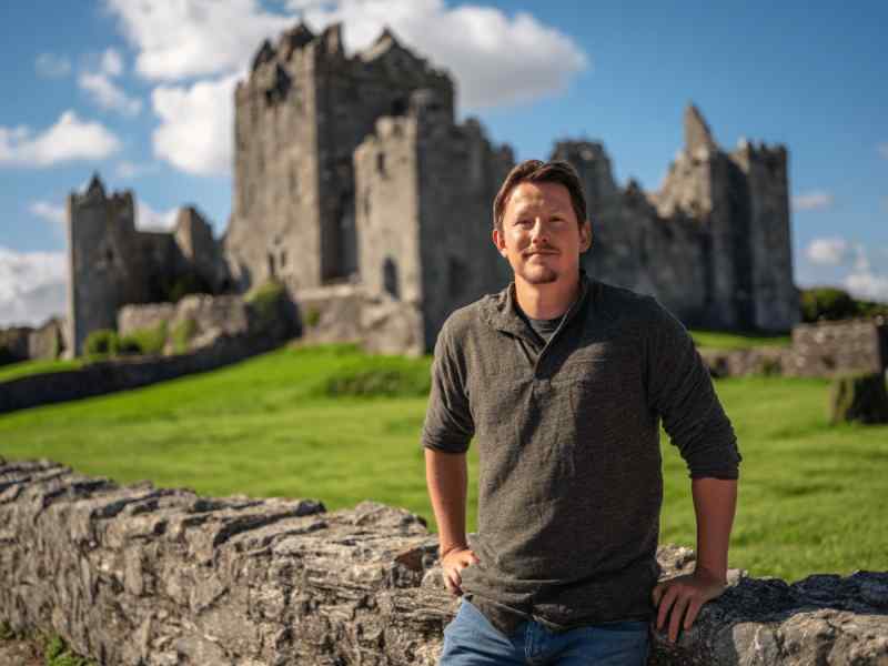 Man standing in front of a medieval stone castle ruin in Ireland on a sunny day, with green grass and historic walls in the background.