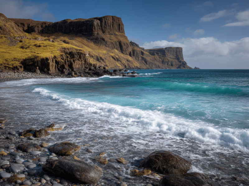 Talisker Bay on the Isle of Skye, Scotland, with waves rolling onto a rocky black sand beach, dramatic sea cliffs rising in the distance, and turquoise waters.