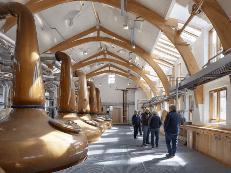Visitors touring a whisky distillery on the Isle of Skye, Scotland, with large copper stills and arched wooden beams inside a bright, modern distilling hall.