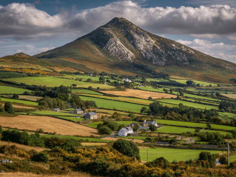 Scenic view of The Great Sugarloaf Mountain rising above green fields and small cottages in County Wicklow, Ireland, with dramatic slopes and a partly cloudy sky.