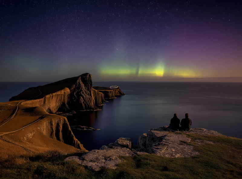 Two people sitting on a cliff overlooking Neist Point Lighthouse on the Isle of Skye, Scotland, with the Northern Lights illuminating the night sky above the Atlantic Ocean.