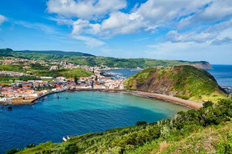 Coastal town with colorful houses by a blue bay, surrounded by lush green hills under a partly cloudy sky.