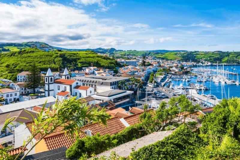 Coastal town with red-roofed buildings, lush hills, and a marina filled with boats under a blue sky with scattered clouds.