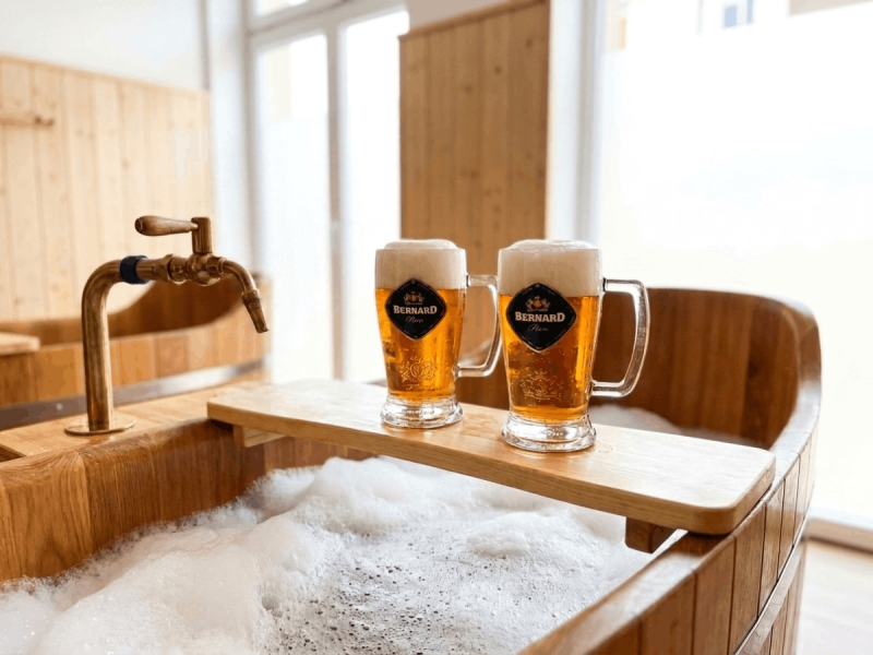 Two mugs of Bernard Unfiltered beer resting on a wooden beer spa tub filled with foam in Prague.