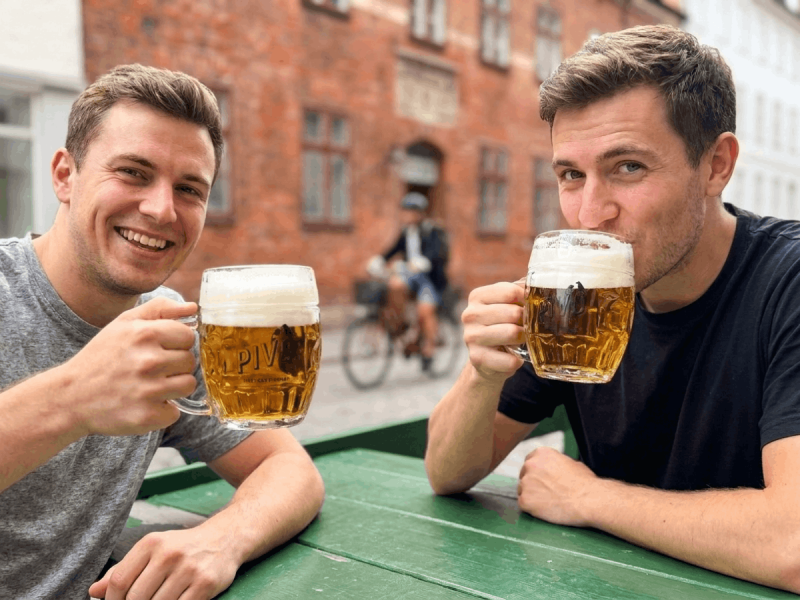 Two men holding mugs of Únětické Pivo at an outdoor cafe table in Prague.