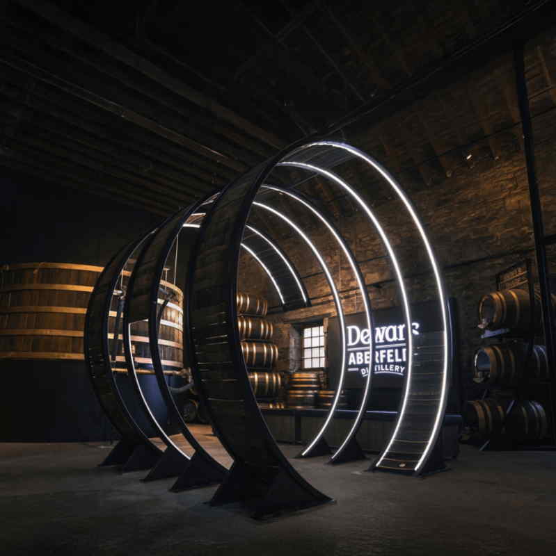 Interior of Dewar’s Aberfeldy Distillery in Perthshire showing a glowing LED barrel-hoop installation, stacked whisky casks, stone walls, and a branded sign in a dim warehouse space.