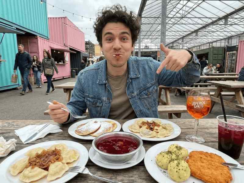 Man pointing at a spread of Czech dumplings, potato pancakes, schnitzel, berry soup, and Aperol Spritz at Manifesto Market, Prague.