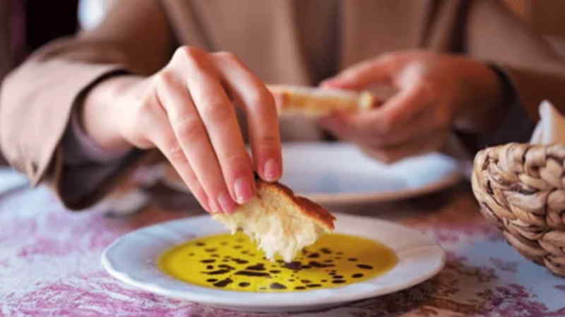 A woman enjoys a piece of bread dipped in olive oil, seated at a table with a relaxed expression.