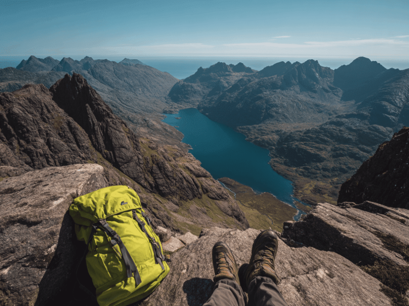 Hiker resting on a rocky ledge in the Cuillin Mountains on the Isle of Skye, Scotland, with boots and backpack in the foreground, overlooking deep blue Loch Coruisk surrounded by dramatic mountain peaks.