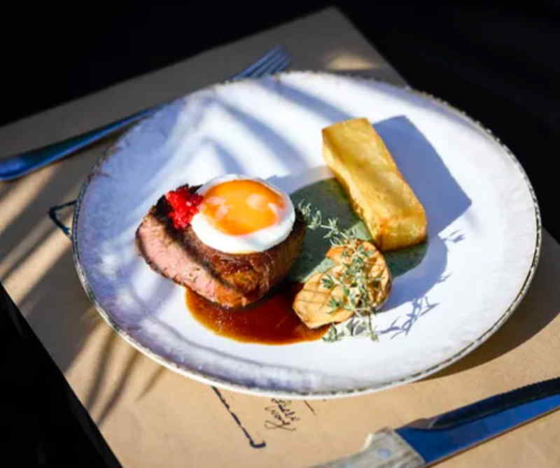 A dish with a grilled steak, seasoned potatoes, and a fried egg on top.