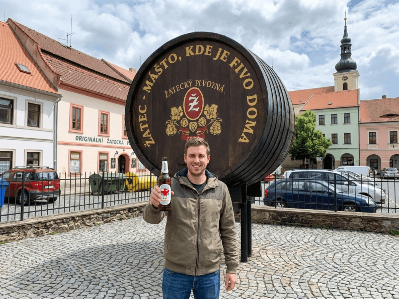 Man holding a bottle of Žatecký Pivovar Pšeničné in front of a giant beer barrel monument in Žatec, Czech Republic.