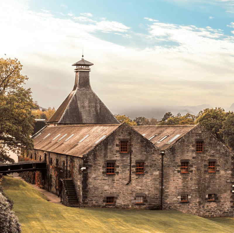 Stone buildings of Glenturret Distillery in Crieff, Perthshire, Scotland featuring a tall pagoda-style kiln roof, red-framed windows and a sloping green lawn beneath a soft, cloudy sky.