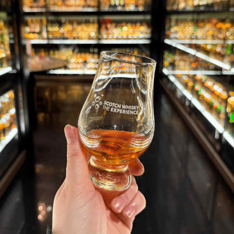 Close-up of a hand holding a Glencairn glass of Scotch whisky inside The Scotch Whisky Experience in Edinburgh, with illuminated shelves of whisky bottles in the background.