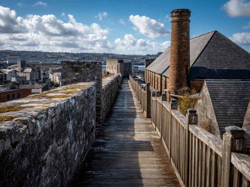 Walkway along the stone ramparts of Elizabeth Fort in Cork City, with historic buildings, brick chimney, and city views stretching into the distance.