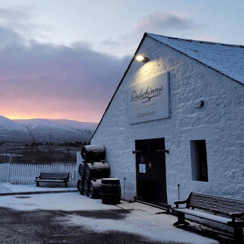 Snow-dusted Dalwhinnie Distillery visitor centre at sunset in the Scottish Highlands, with stacked whisky barrels, benches, white picket fence, and mountain backdrop.