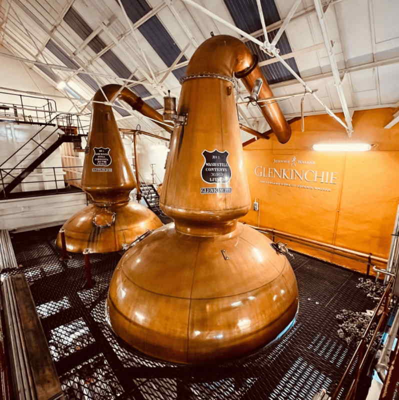 Interior of Glenkinchie Distillery in East Lothian showing large polished copper pot stills under a white trussed roof, metal walkways and pipes, with an orange wall bearing Glenkinchie signage.