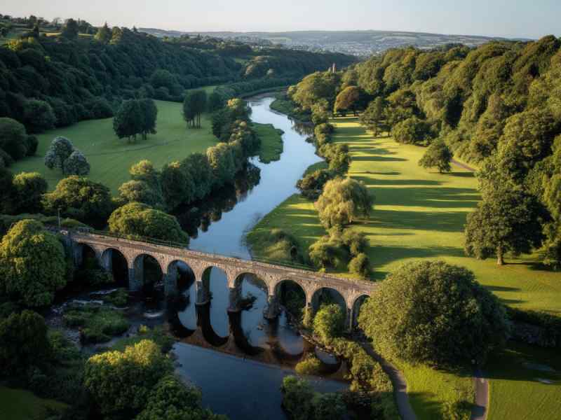 People walking and relaxing along the River Lee in Ballincollig Regional Park, surrounded by open green space, trees, and riverside paths.
