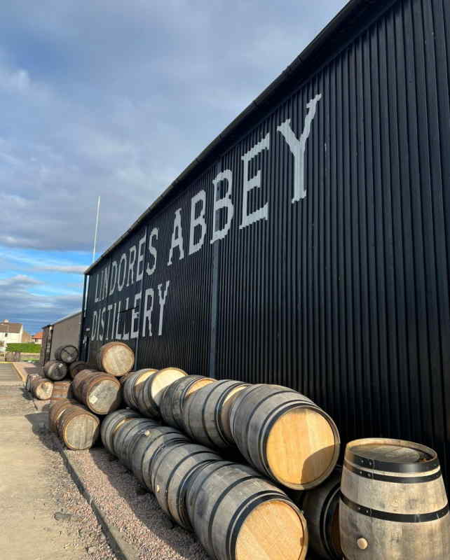 Stacked oak whisky barrels beside a black corrugated warehouse painted “LINDORES ABBEY DISTILLERY” at Lindores Abbey Distillery in Newburgh, Fife, under a bright partly cloudy sky.
