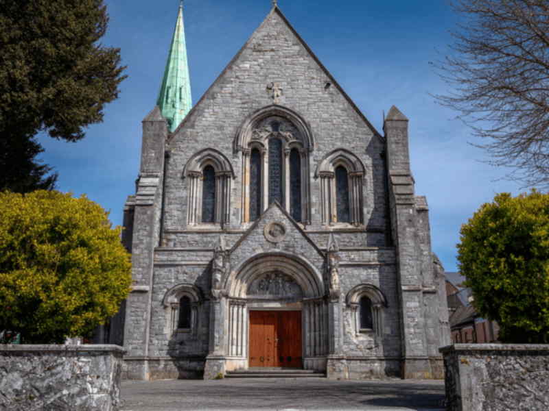Front view of a church in Finglas, Dublin, featuring its stone façade, Gothic-style arched doorway, tall spire, and intricate stained-glass windows under a clear blue sky.