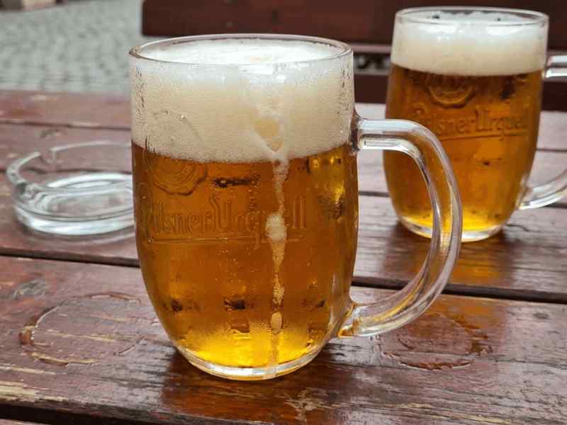 Two cold Pilsner Urquell beer mugs with frothy heads on a wooden pub table in Prague, Czech Republic.