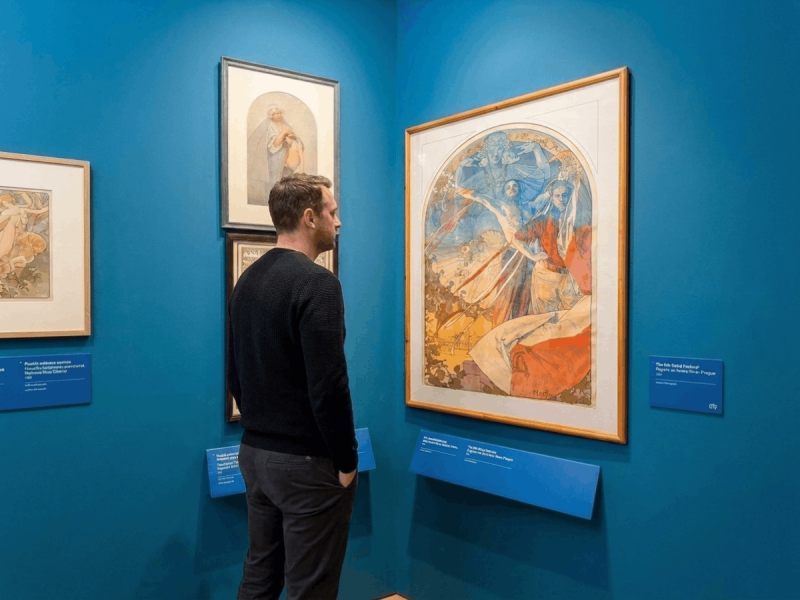Man viewing framed Art Nouveau illustrations on a blue gallery wall at the Mucha Museum, a top pick among Prague museums.