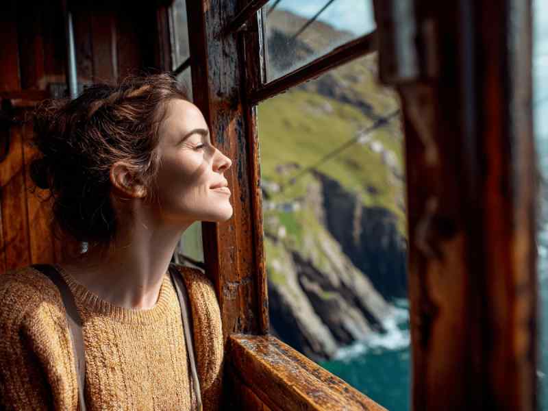 Woman looking out a wooden window toward a rugged coastline and ocean cliffs.