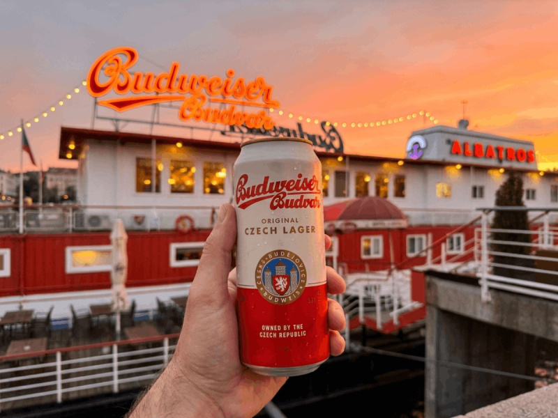 Hand holding a can of Budweiser Budvar Czech lager at sunset in front of the Albatros boat bar in Prague.