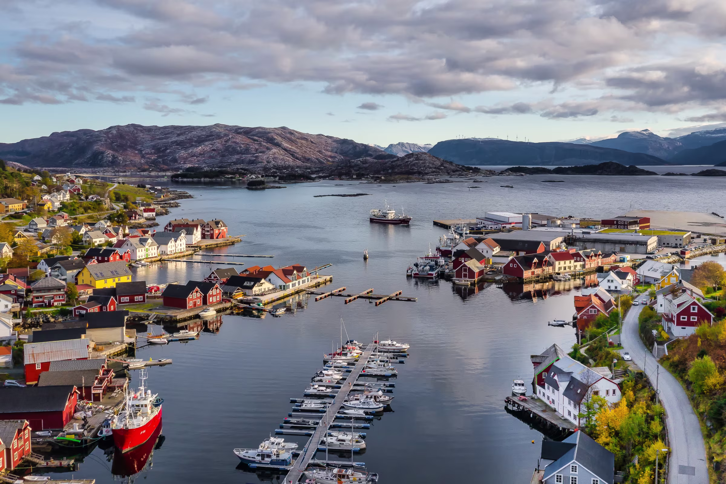 En havn med flere båter og hus rundt havnen. Øyer og fjell i bakgrunnen.
