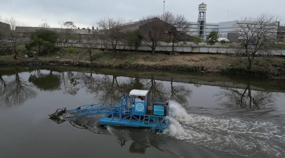 A slow-moving catamaran operated by the Autoridad de Cuenca Matanza Riacheulo (ACUMAR) turns a corner near Villas 21-24 in the Autonomous City of Buenos Aires as it sifts garbage from the river into its collection bucket, 2023.