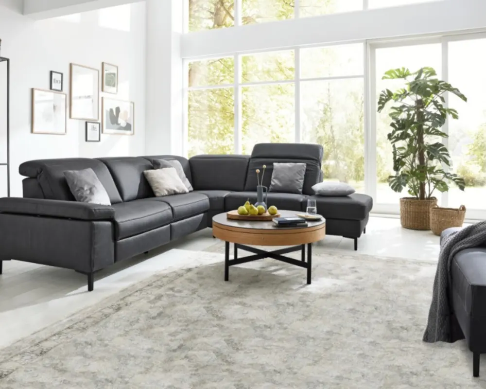 Modernes Wohnzimmer mit grauem Ecksofa, hellen Akzenten und Panoramablick durch bodentiefe Fenster. Wohnzimmer mit grauem Ecksofa, Couchtisch aus Holz und Metall, großem Fenster, Wandbildern und Teppich in Naturtönen