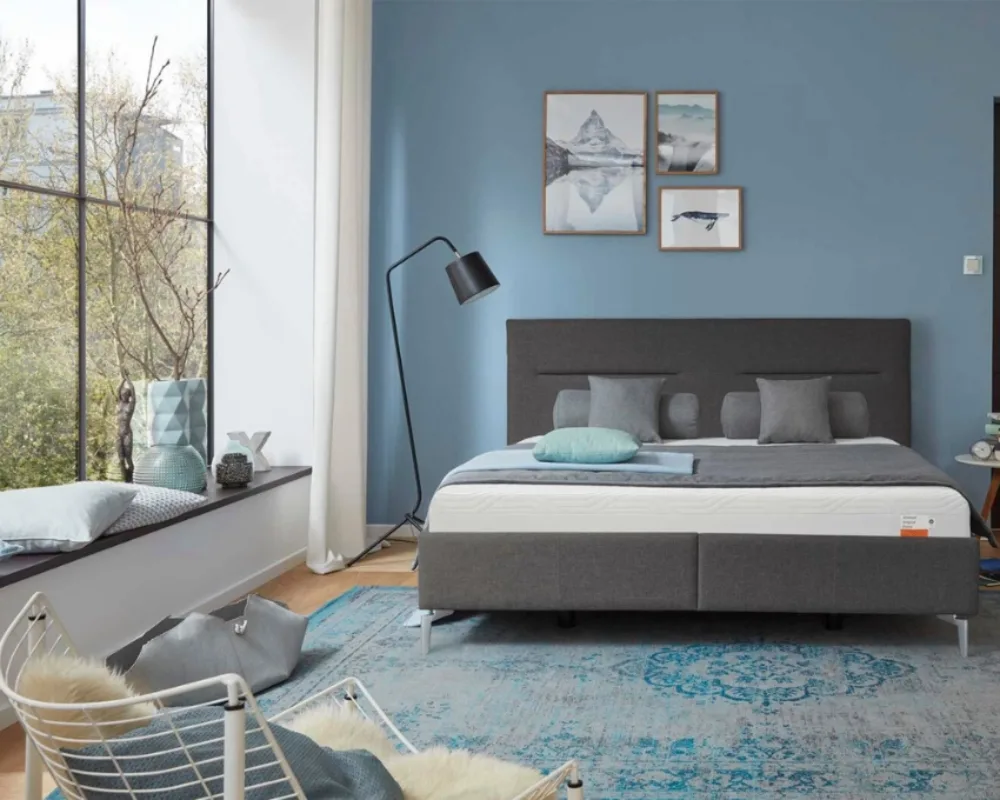 Tempur Boxspringbett in urbanem Schlafzimmer Graues Tempur Bett vor großen Fenstern mit moderner Wanddekoration und natürlichem Licht