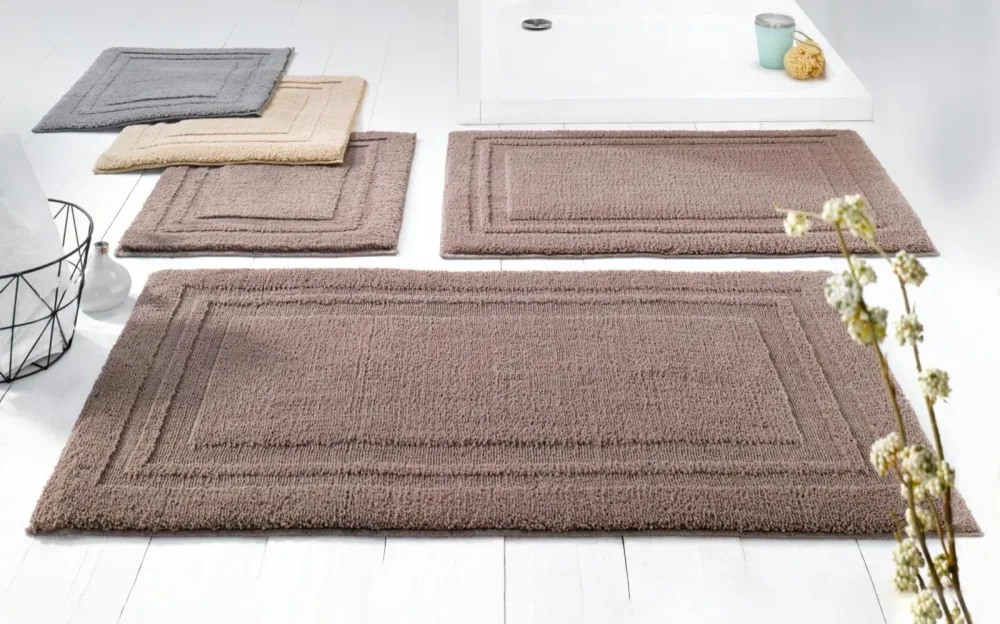 Badematten-Set in Naturtönen vor moderner Dusche mit Badzubehör und Blumen Vier Badematten in Grau, Beige und Braun vor weißer Dusche mit Schwamm, Behälter und Blumenstrauß auf weißem Boden.