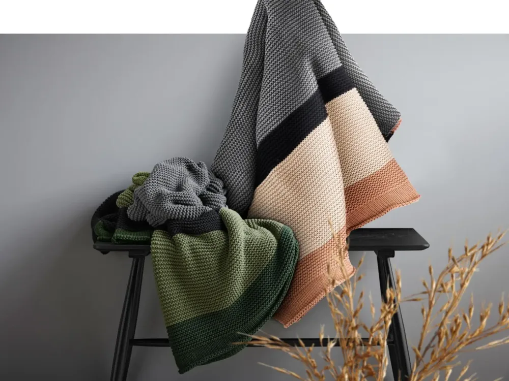 Strickdecken in Erdtönen auf schwarzer Bank vor grauer Wand Zwei grob gestrickte Decken in Naturtönen, Beige-Rosé und Grün-Schwarz, dekorativ über schwarze Holzbank gelegt